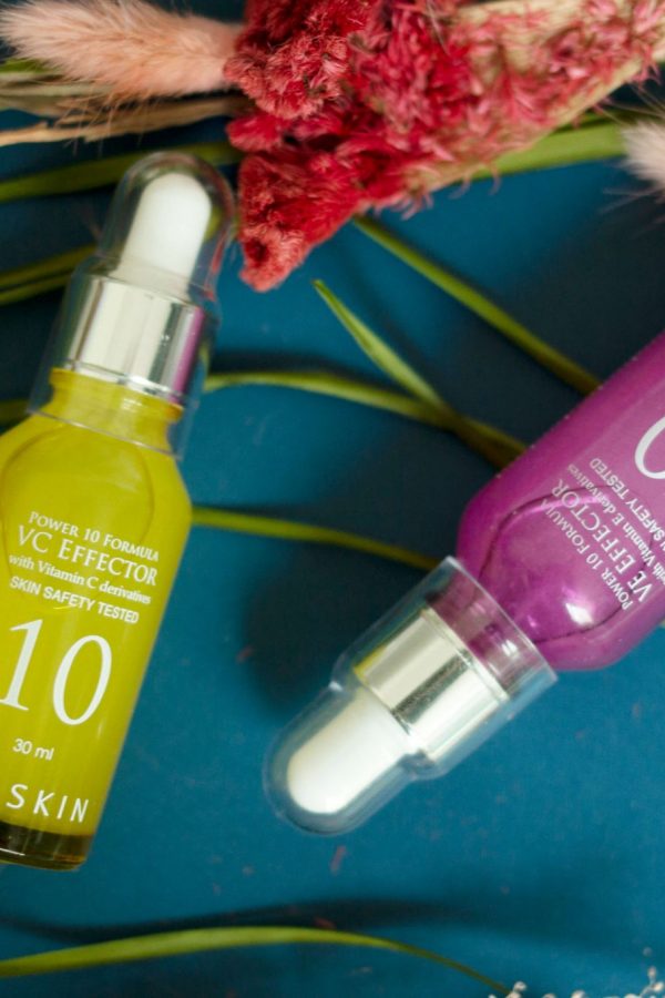 overhead shot of two brightly coloured glass bottles from the korean beauty brand It'S SKIN on top of flowers in a flatlay style photo. The left bottle is bright yellow and contains the Power 10 Formula VC Effector, the right hand bottle is a deep purple, and contains the Power 10 Formula VE Effector