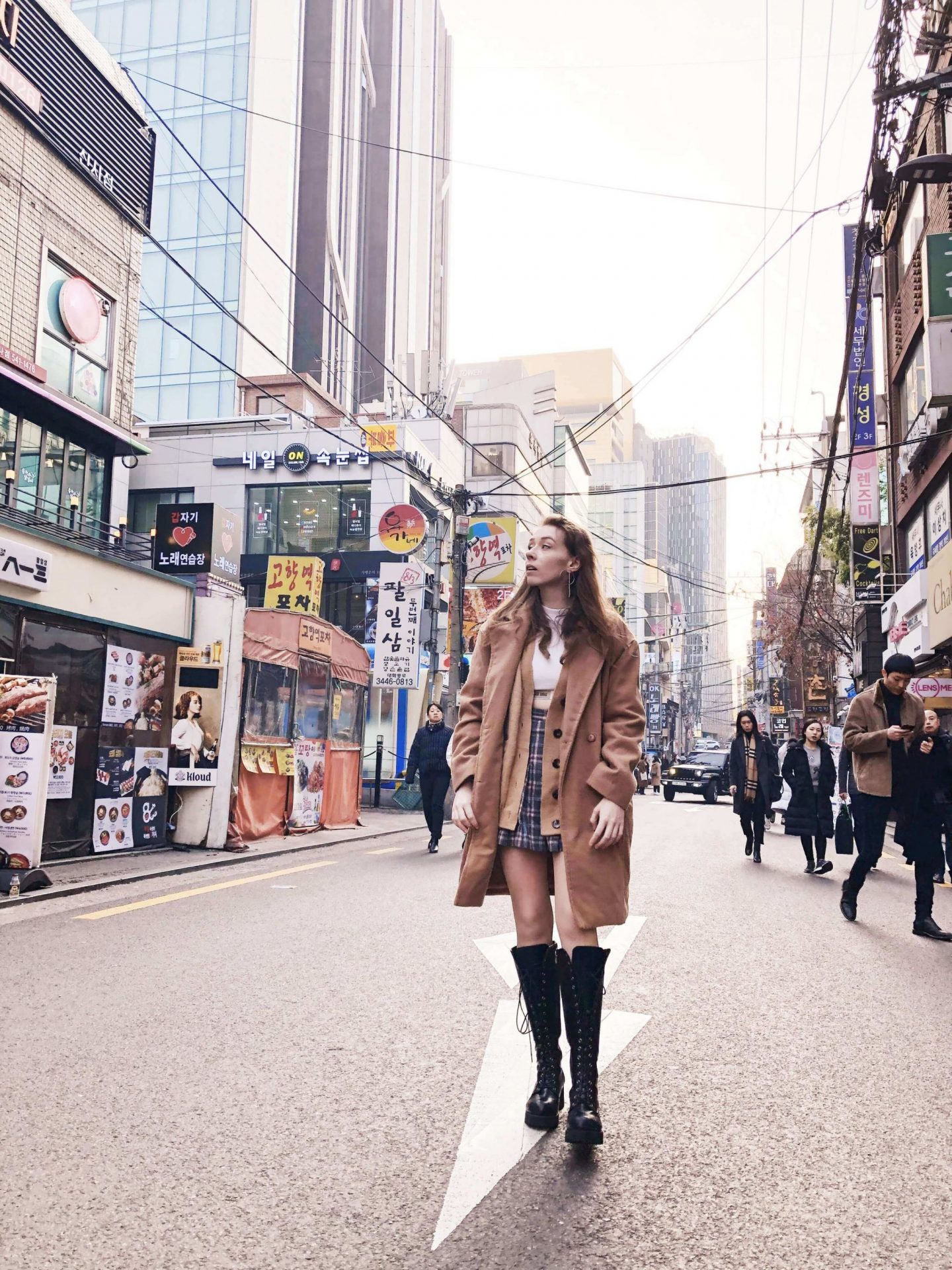 An image of Fii standing in the middle of a road with tall buildings and people either side. She is wearing a knee length brown coat with the buttons undone, so that you can see some of the clothes she wears underneath: a check pattern skirt and a brown cardigan. She is also wearing knee high black lace up boots, and is looking off to one side so you can see the long earrings. 
