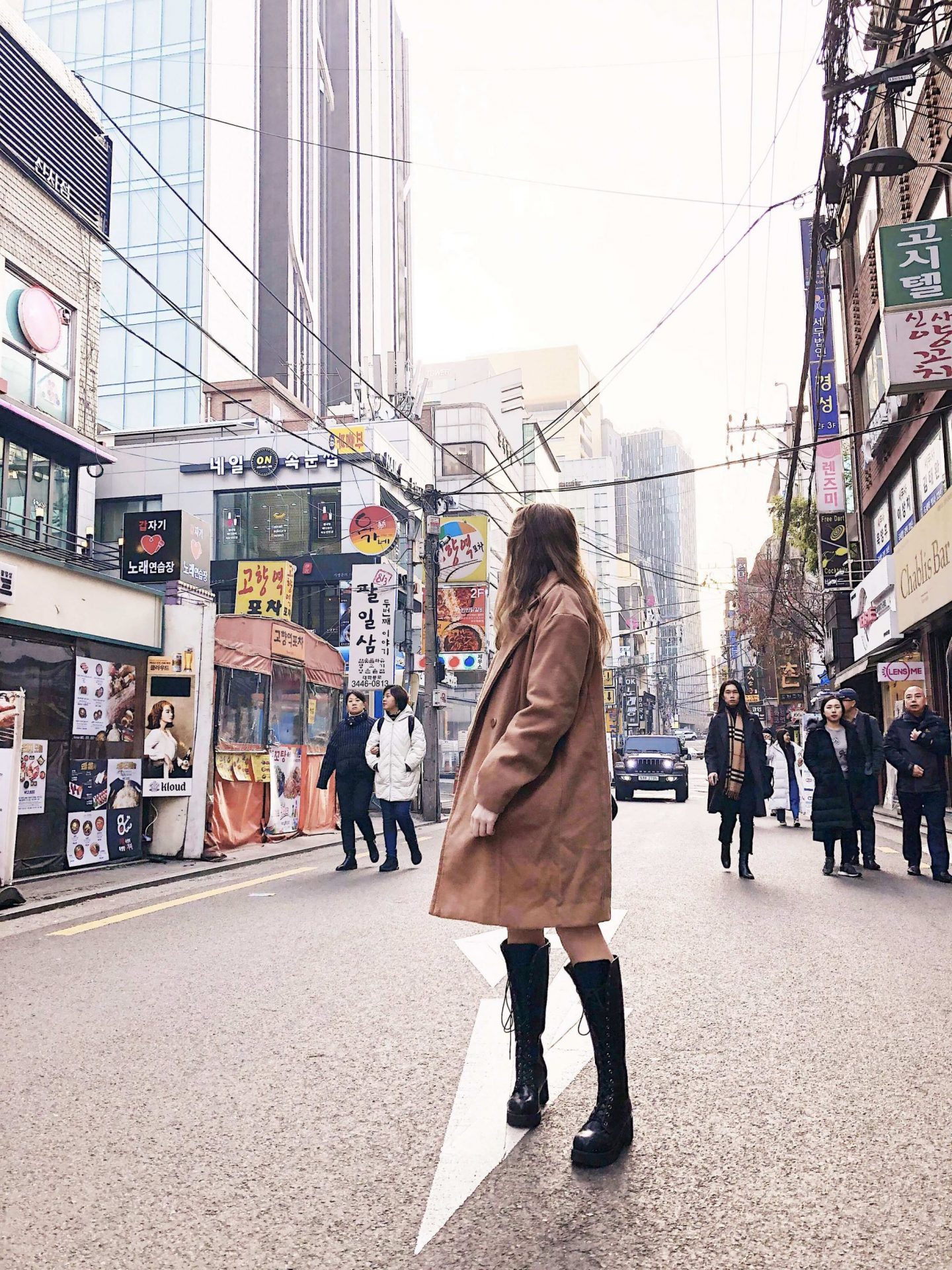 image of fii standing in the middle of a road looking backwards, wearing a knee-length camel coat, knee high black boots. There are tall buildings either side with Korean signs on them