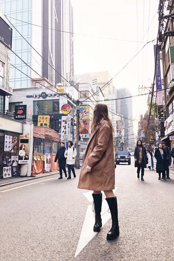 image of fii standing in the middle of a road looking backwards, wearing a knee-length camel coat, knee high black boots. There are tall buildings either side with Korean signs on them