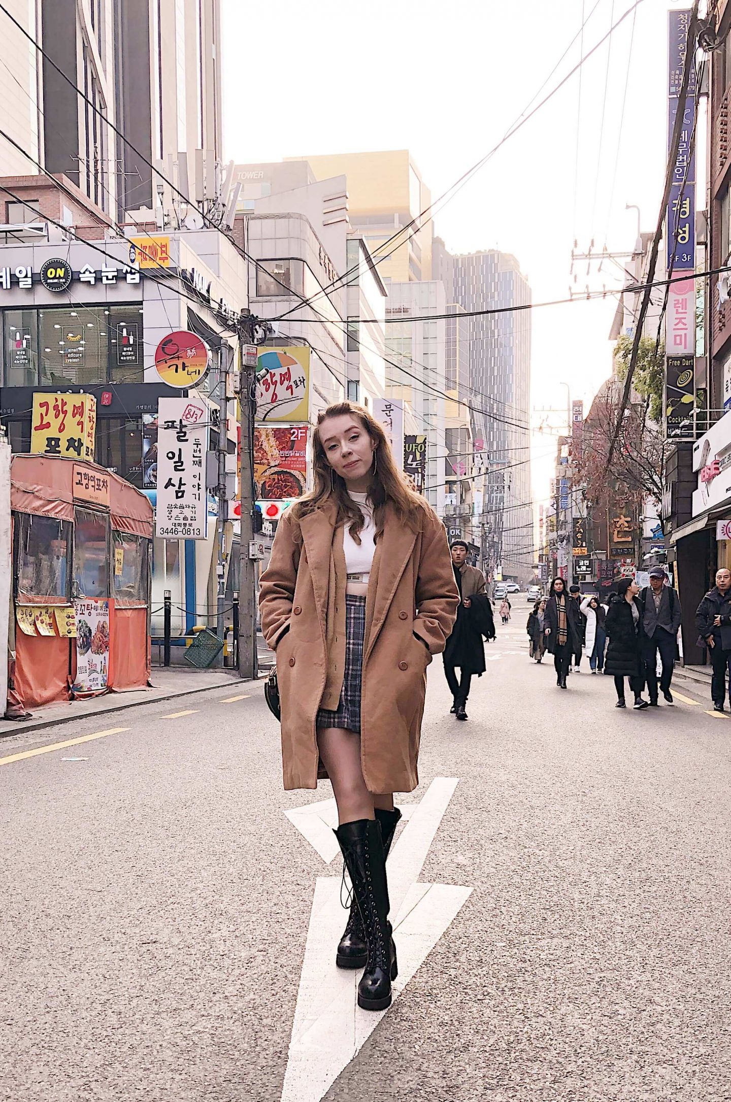 Fii is standing in the middle of the road facing the camera, with tall buildings either side and some people behind. She is wearing the same outfit as above and almost smiling at the camera. 